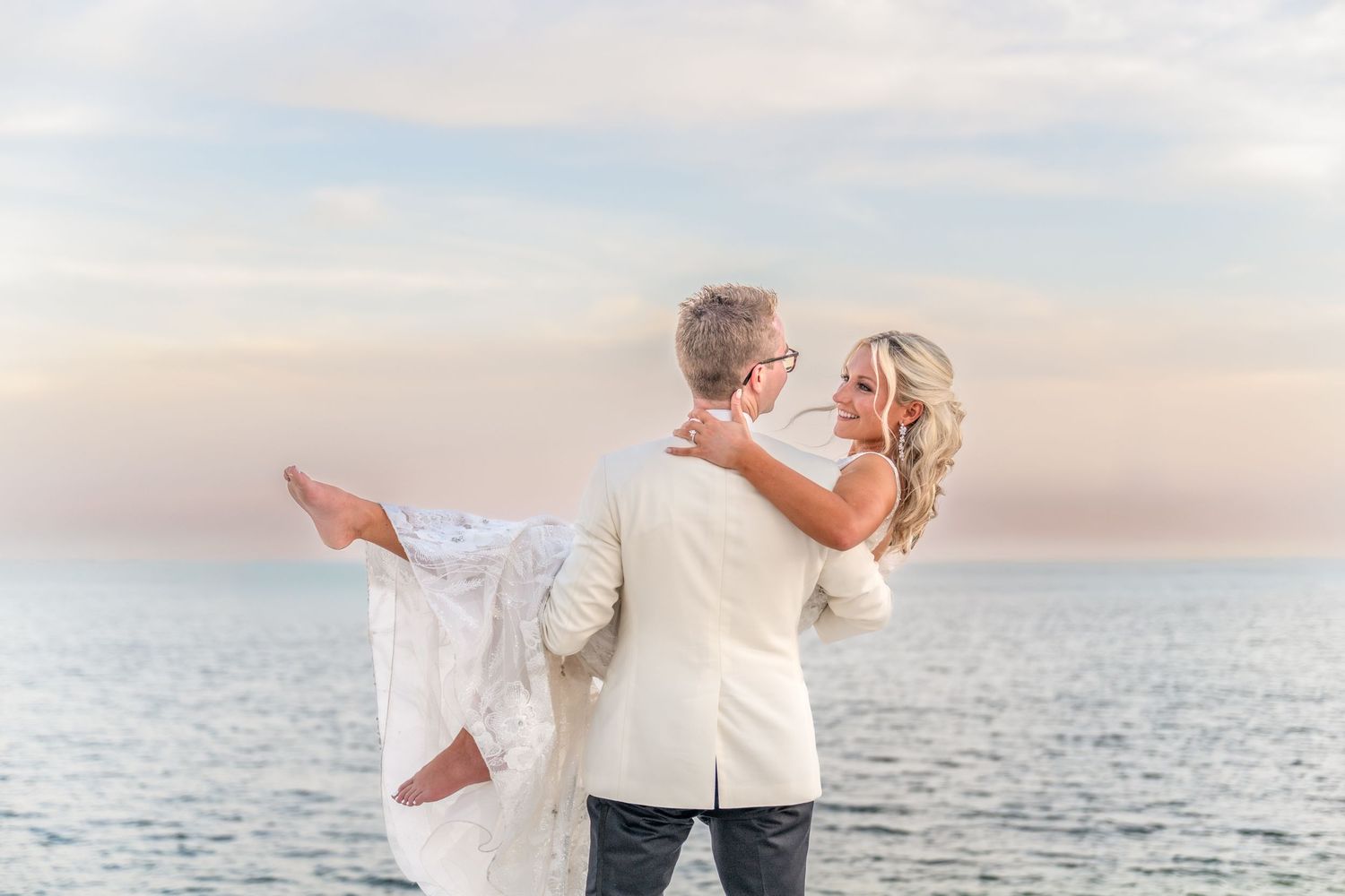 Groom in white suit carries bride in lace dress on a serene beach at sunset, sharing a romantic moment.
