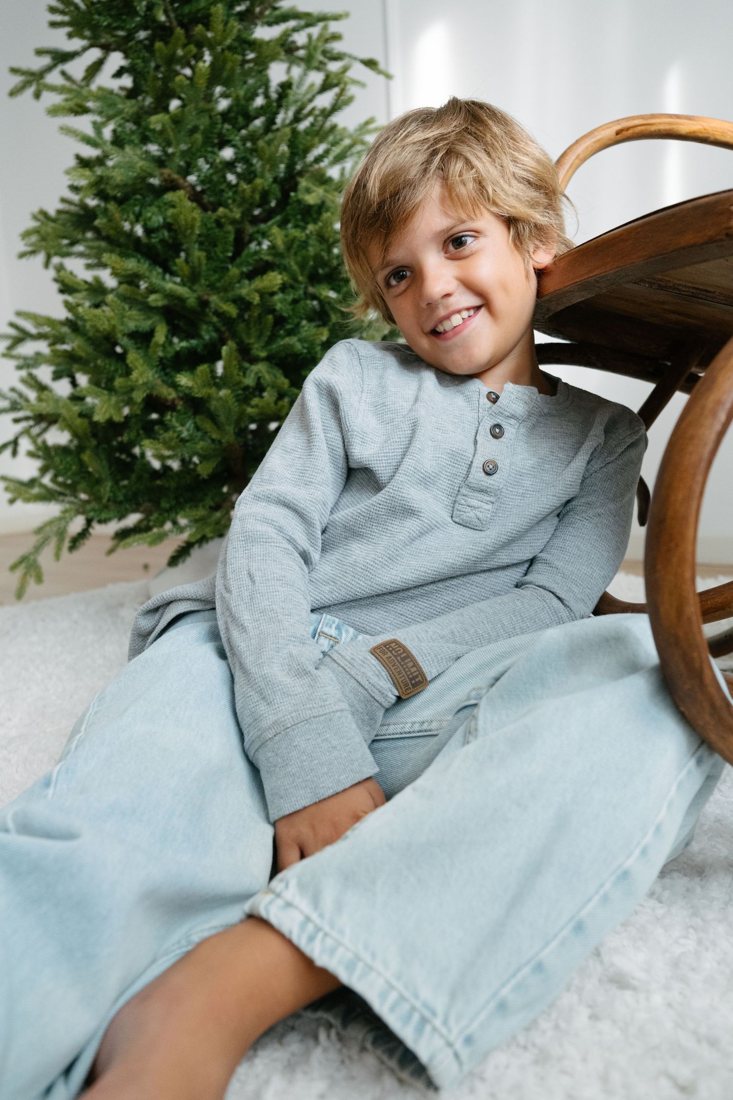 Smiling blond boy in gray henley and jeans sitting on floor beside wooden chair and Christmas tree.