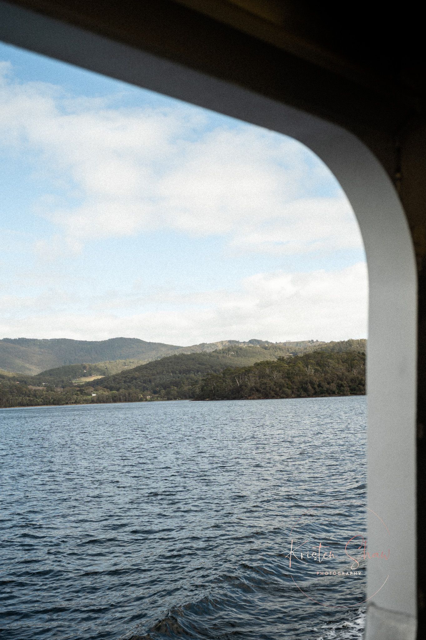 View of forested hills and calm blue water framed by a boat window under a partly cloudy sky.