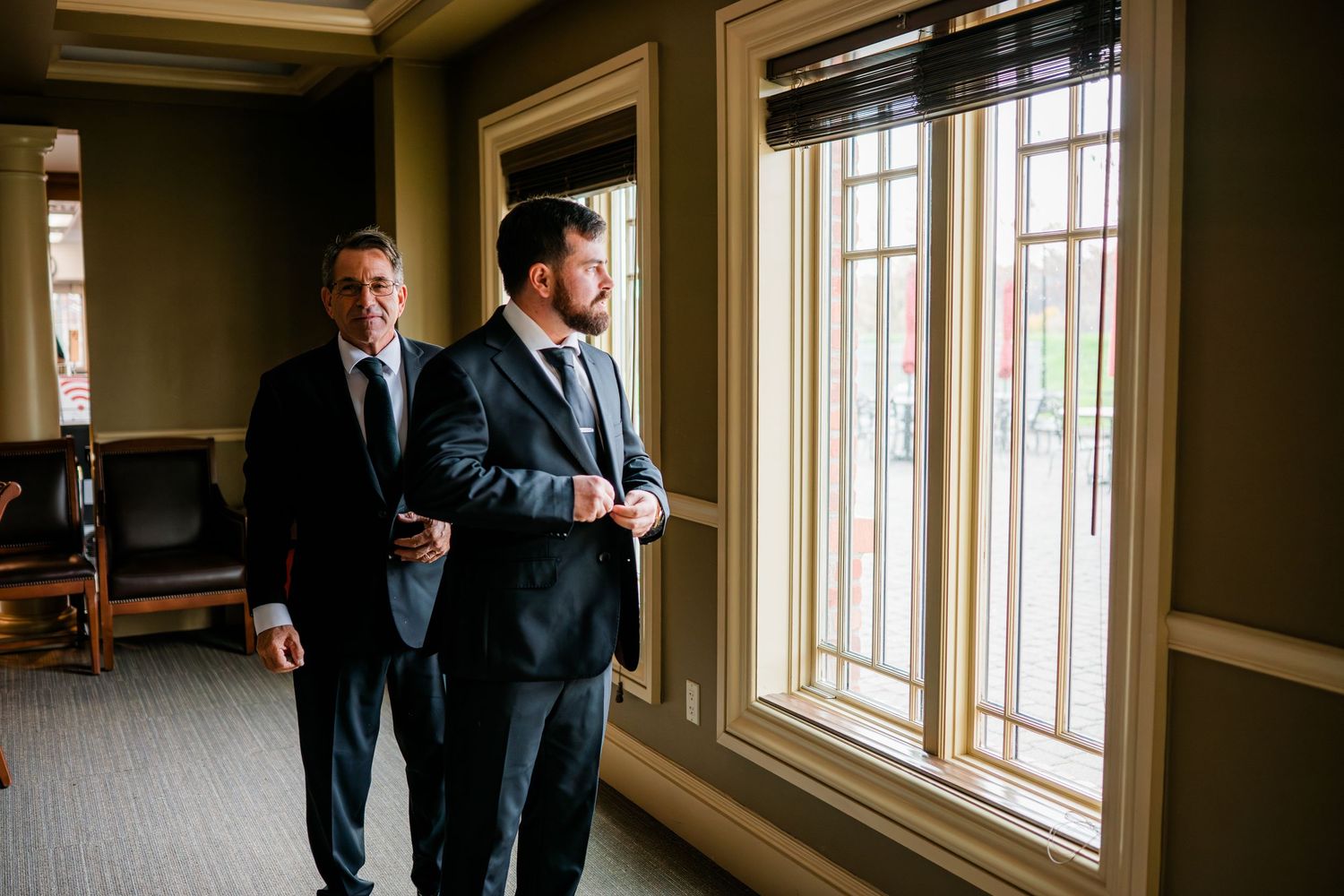 Two men in dark suits stand by elegant wooden-framed windows in a formal interior setting at Great River Golf Course, in Orange, CT