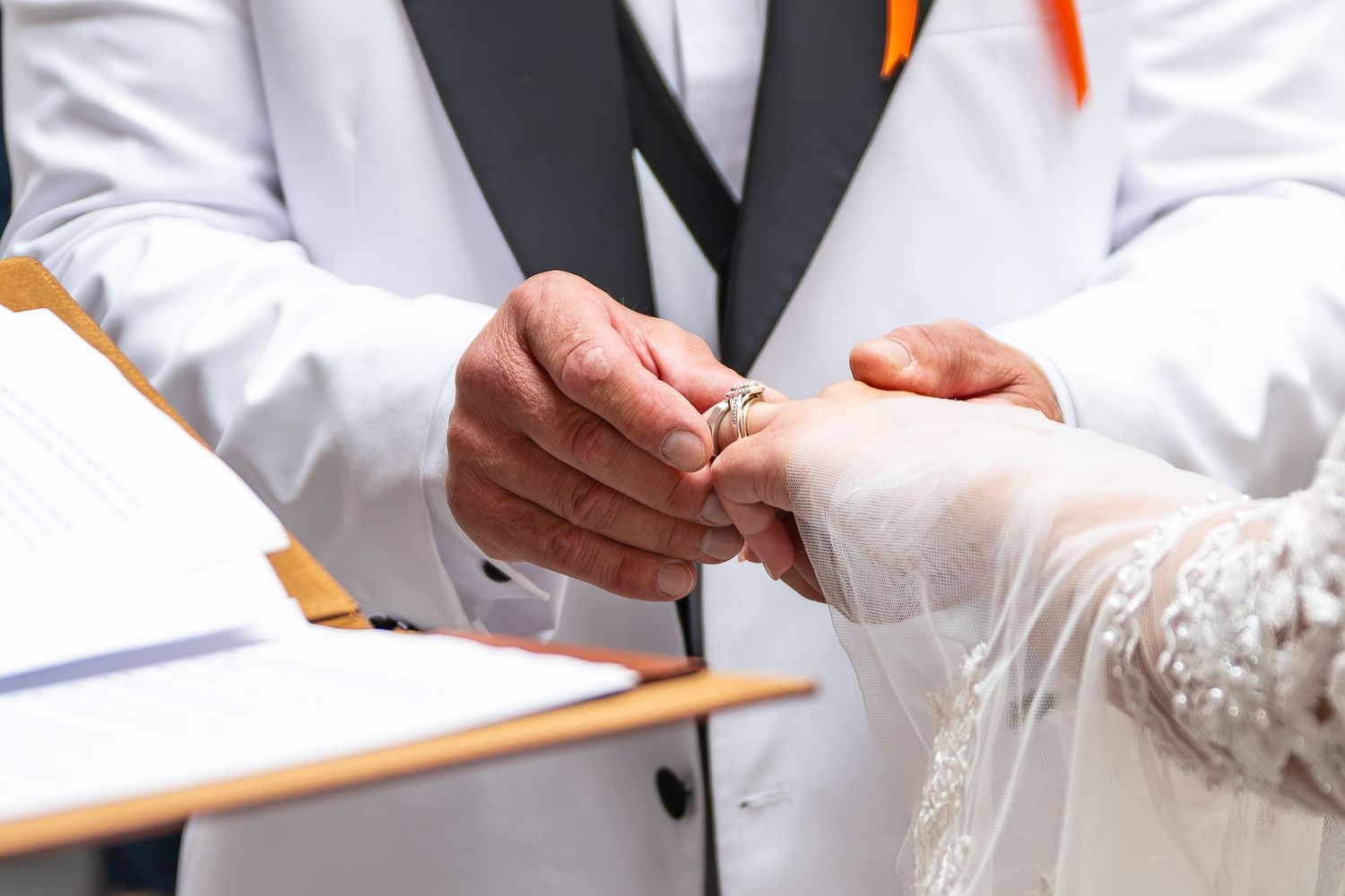Groom placing a wedding ring on bride's finger during ceremony, with marriage register and white suit visible.Yorkshire, Sheffield, UK