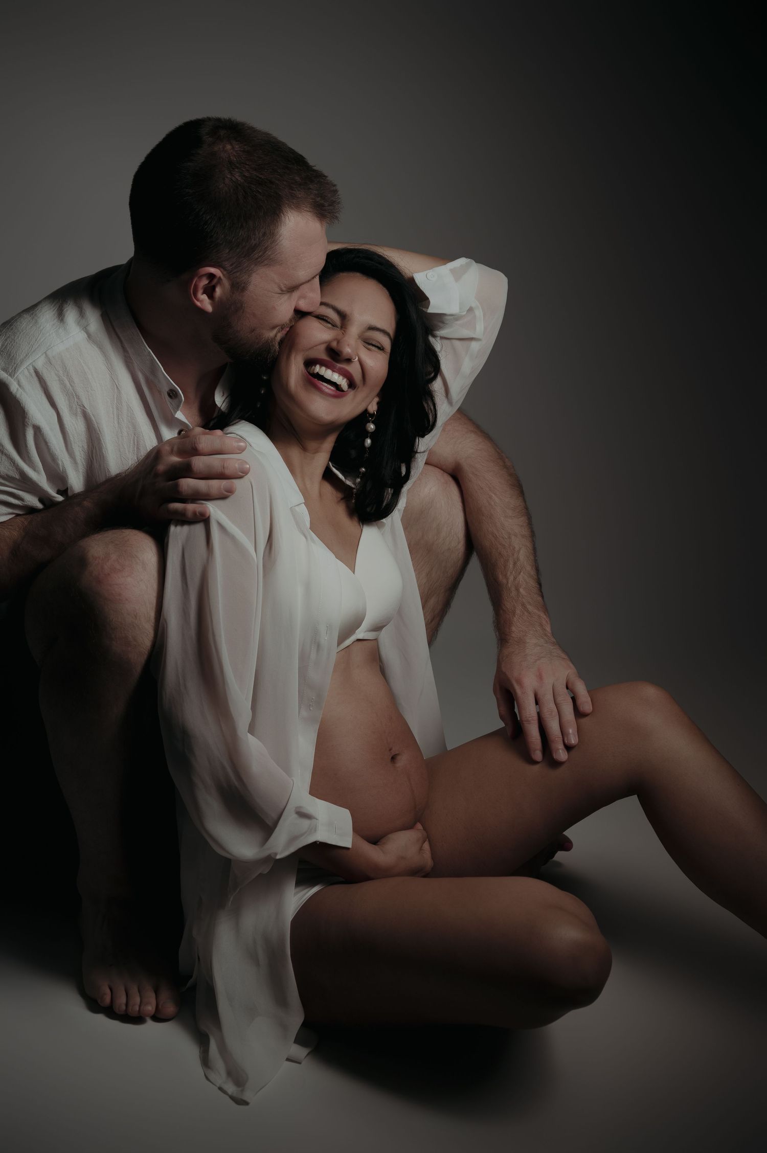 A laughing pregnant woman in a white sheer robe sits on the floor as her partner kisses her cheek in a studio.