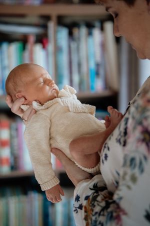 A sleeping newborn in a cream knit sweater cradled gently in a mother's arms near bookshelves.