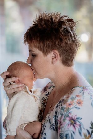 A mother with short brown hair tenderly kisses her newborn baby dressed in a cream knit outfit during in home newborn photos.