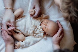 Newborn baby sleeping peacefully on white blanket while parents gently hold tiny hands and feet.
