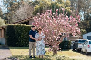 Couple standing together under a blooming pink magnolia tree in front of a suburban house on a sunny day.