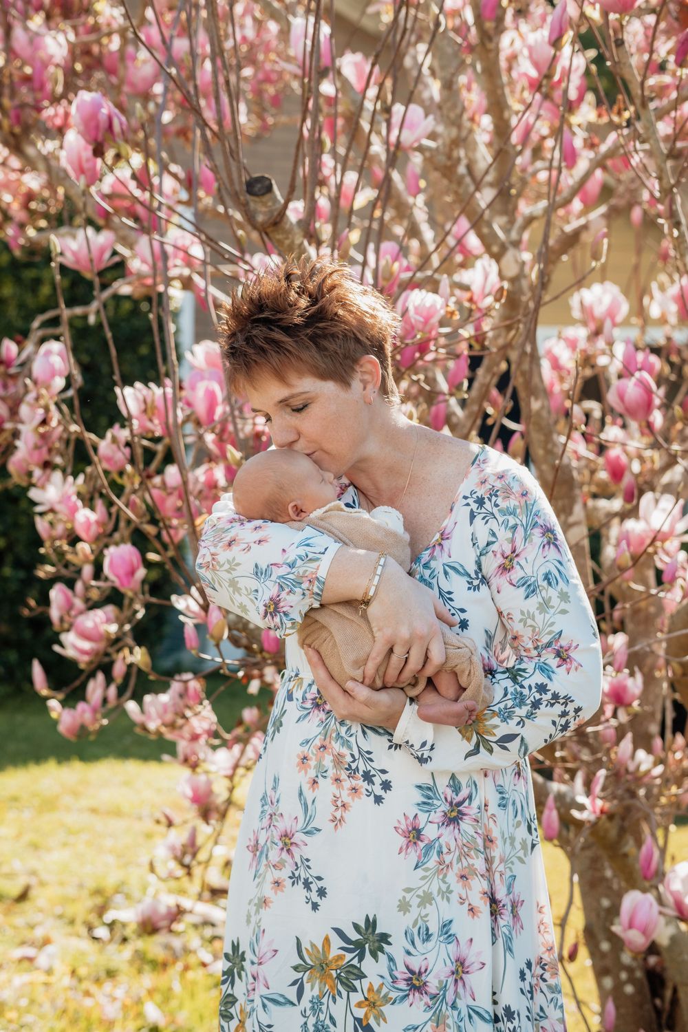 A woman in a floral dress tenderly kissing her newborn baby while standing beside a blooming pink magnolia tree outdoors.