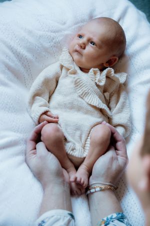 A newborn baby in a cream knit romper lies on a white blanket, cradled gently by a mother's hands.