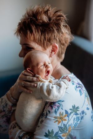 Woman in floral top tenderly holds yawning newborn baby close to her chest during in home newborn photos.
