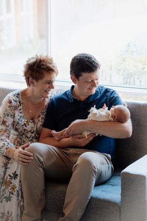 A couple sitting on a gray sofa smiling at their newborn baby held gently in the fathers arms near a bright window.