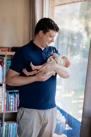 Father in navy polo shirt lovingly cradles newborn baby near bright window.