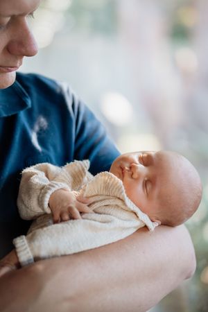 A parent gently cradling a sleeping newborn baby dressed in a cream knit outfit, sharing a tender moment.