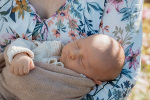 Sleeping newborn baby cradled in arms wearing a knit outfit, held by a person in a floral top outdoors.