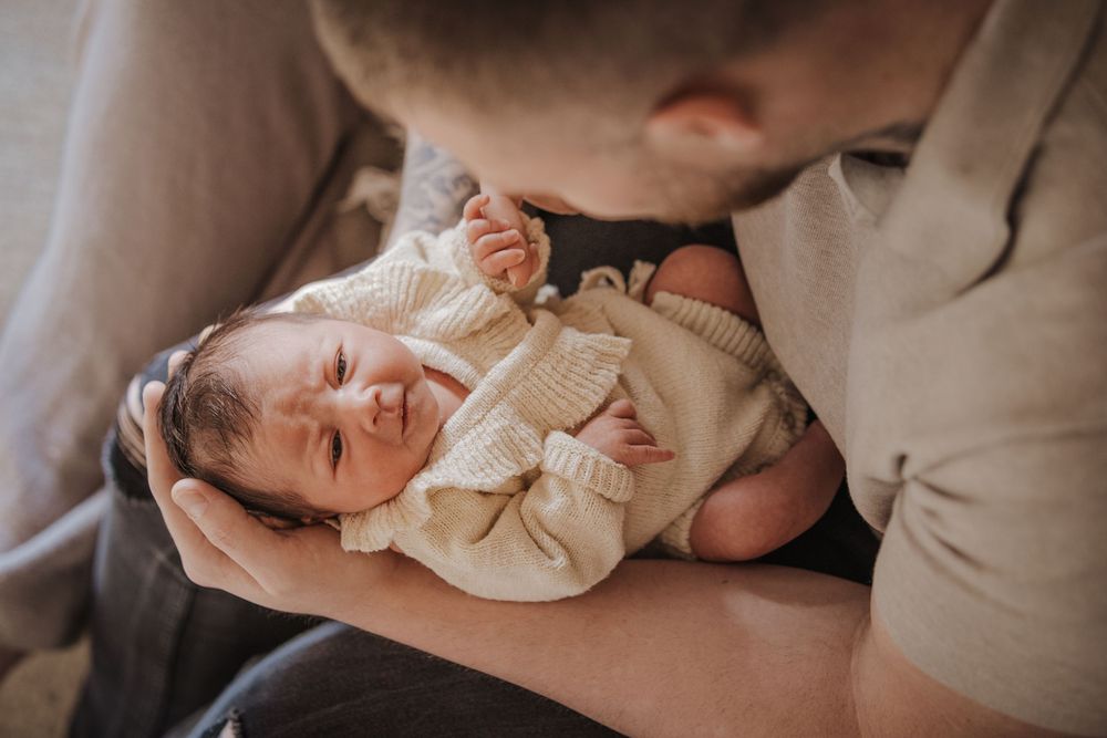 A father tenderly kisses his newborn baby dressed in a cream knit outfit while cradling them gently.