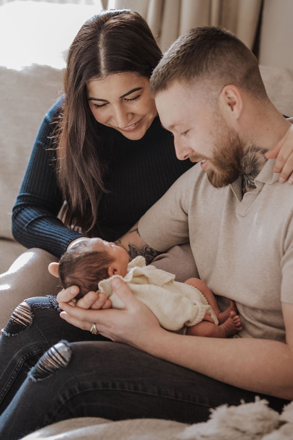 Young couple sitting on couch smiling at their newborn baby wrapped in a white blanket.