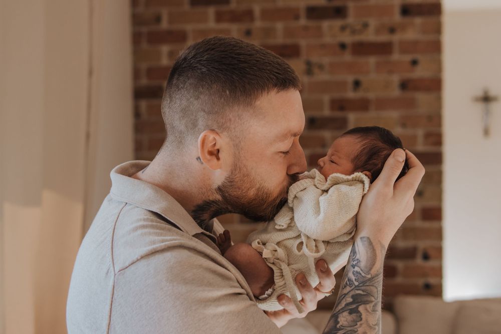A tattooed father tenderly kisses his newborn baby while holding the infant close in a warm home setting.