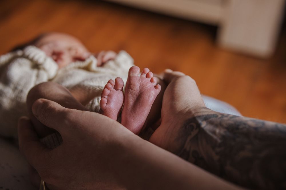 Tattooed parent gently holds tiny newborn baby feet in their hands on a wooden floor background During in home newborn photos in Castle Hill