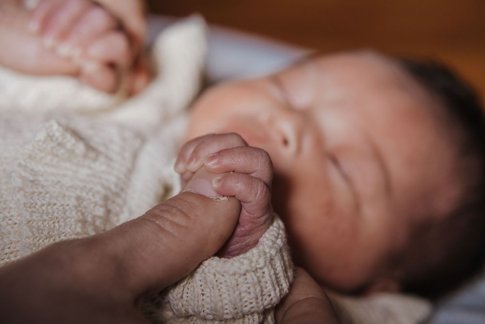Newborn baby sleeping peacefully while gently gripping an adult finger, wrapped in a soft knit blanket.