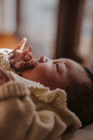 Peaceful newborn baby sleeping wrapped in a cream knit blanket, tiny fingers reaching upward in soft natural light.