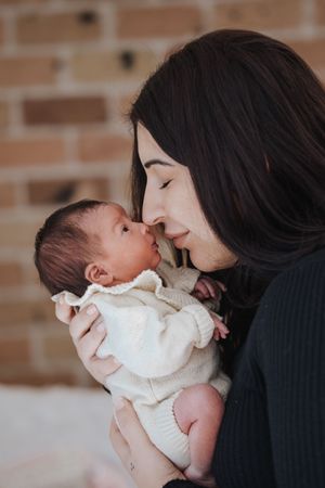 A mother tenderly touches noses with her newborn baby, both dressed warmly, against a brick wall background during in home newborn photos