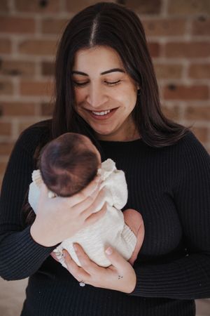 Smiling dark-haired woman in black sweater tenderly holding a newborn baby in white outfit against brick wall.