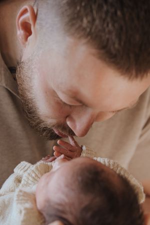 A bearded father tenderly kisses his newborn baby's tiny hand while cradling the infant in a soft knit blanket.
