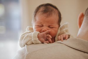 A peaceful newborn baby sleeps soundly in a white knit outfit while resting on a parents shoulder.