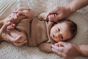 Newborn baby in brown knit outfit lying on white blanket while parents gently hold hands and feet.