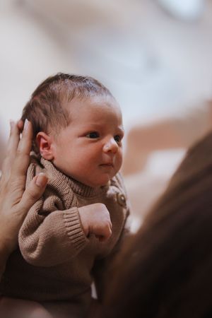 A newborn baby with dark hair wearing a beige knit sweater being cradled in loving hands.