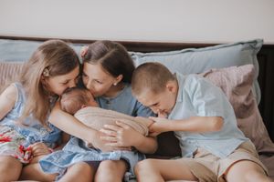 A mother and three children cuddle together on a bed, lovingly admiring a newborn baby.