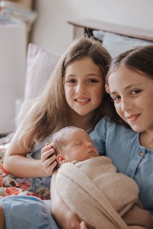 Two young girls smiling while holding a sleeping newborn baby wrapped in a beige swaddle blanket at home in The Ponds.