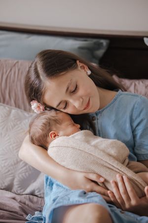 Young mother tenderly holding her sleeping newborn baby wrapped in a soft blanket while sitting on a bed.