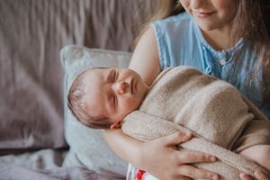 A mother in a blue dress tenderly holds her sleeping newborn baby wrapped in a soft beige blanket.