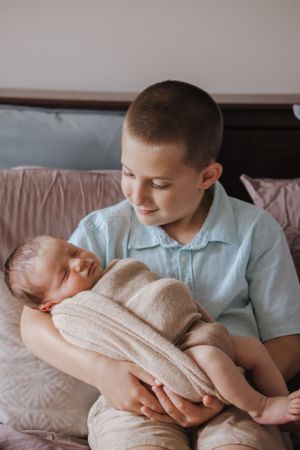 Young boy in blue shirt smiling while gently holding sleeping newborn baby wrapped in beige knit blanket.