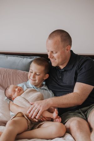 A smiling father and young son sitting on a bed together lovingly holding a sleeping newborn baby.