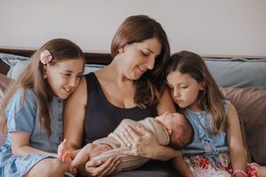 A mother sits on a bed holding her newborn baby, flanked by her two young daughters in blue dresses.