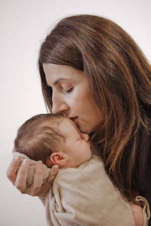 A mother with brown hair tenderly kisses her sleeping newborn baby wrapped in a beige blanket.