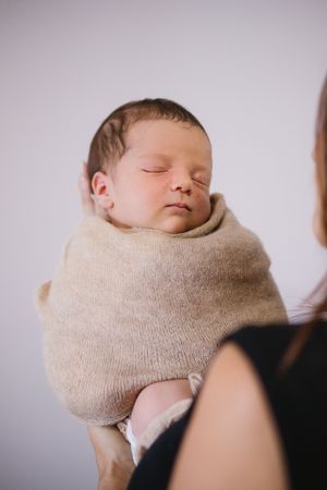 Sleeping newborn baby swaddled in soft beige knit wrap being held by mother against white background at home in The Ponds.