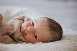 Newborn baby with dark hair lying on a soft white blanket, wearing a brown onesie, with wide curious eyes.