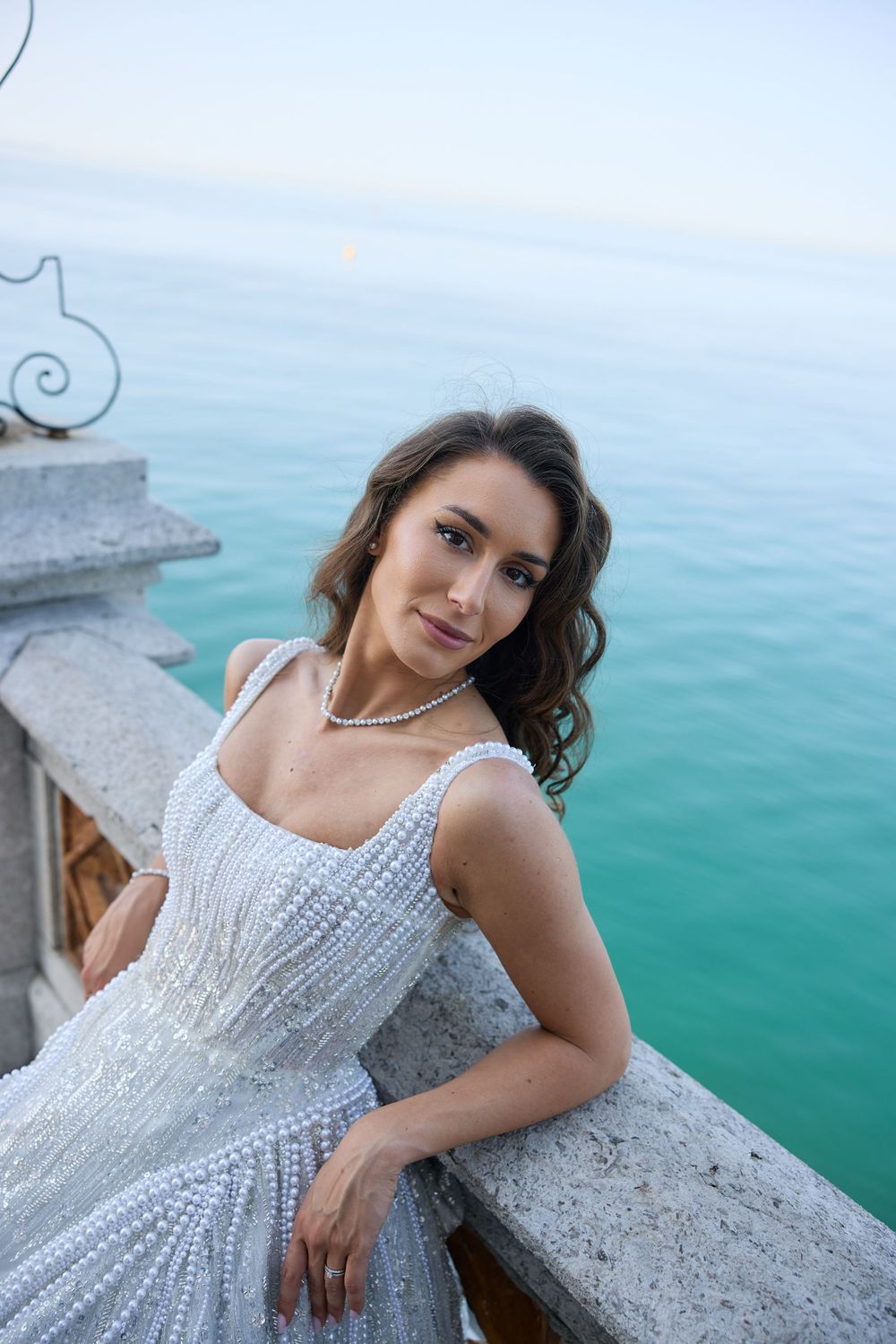 Elegant bride in pearl wedding gown leans on stone balustrade with turquoise sea backdrop.