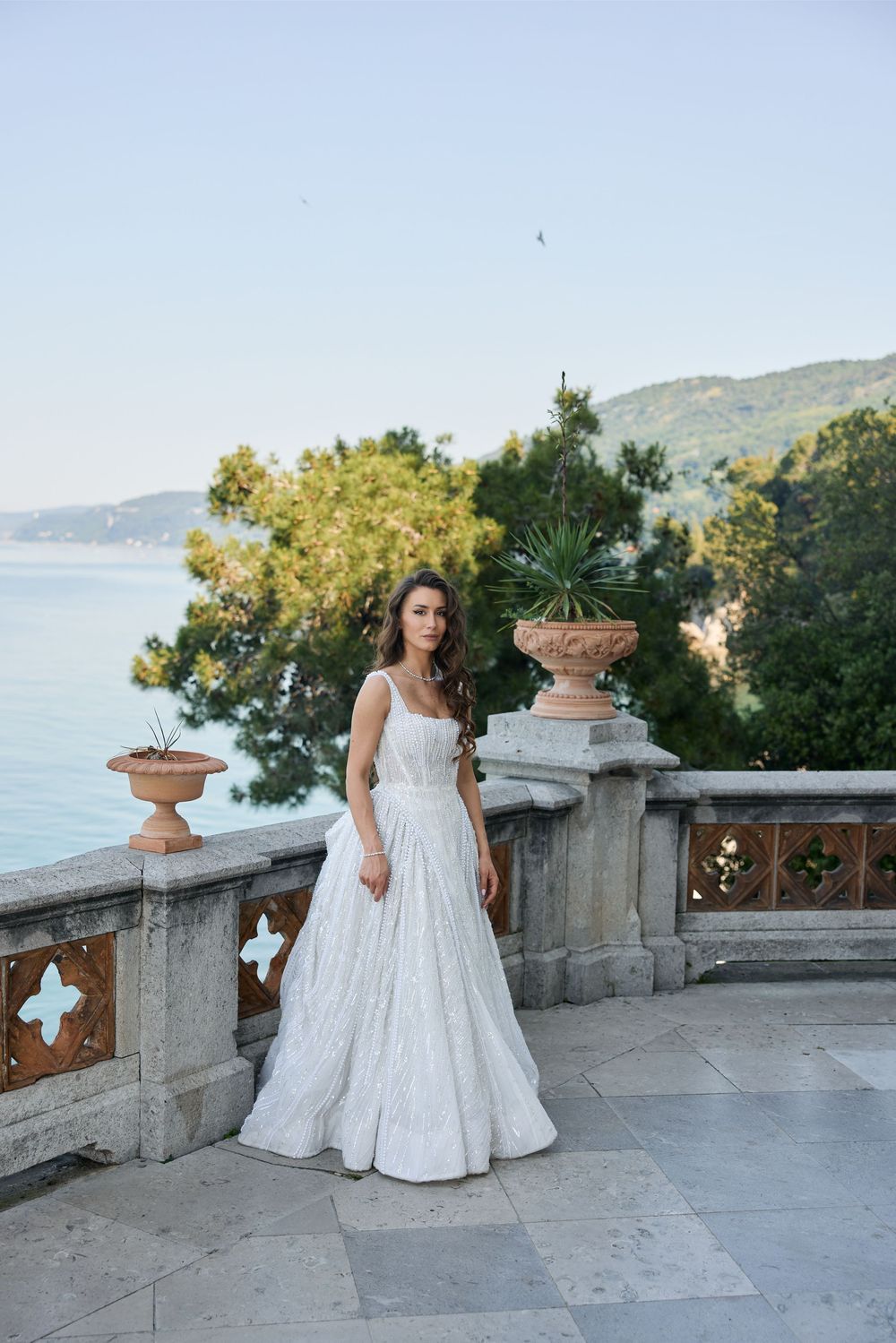 Bride in white ball gown stands on terrace with decorative urns overlooking scenic coastal landscape.