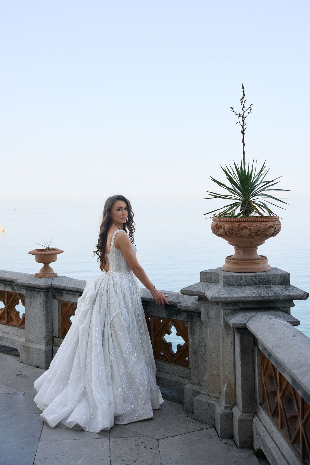 Bride in flowing white ball gown poses on stone terrace overlooking calm ocean.