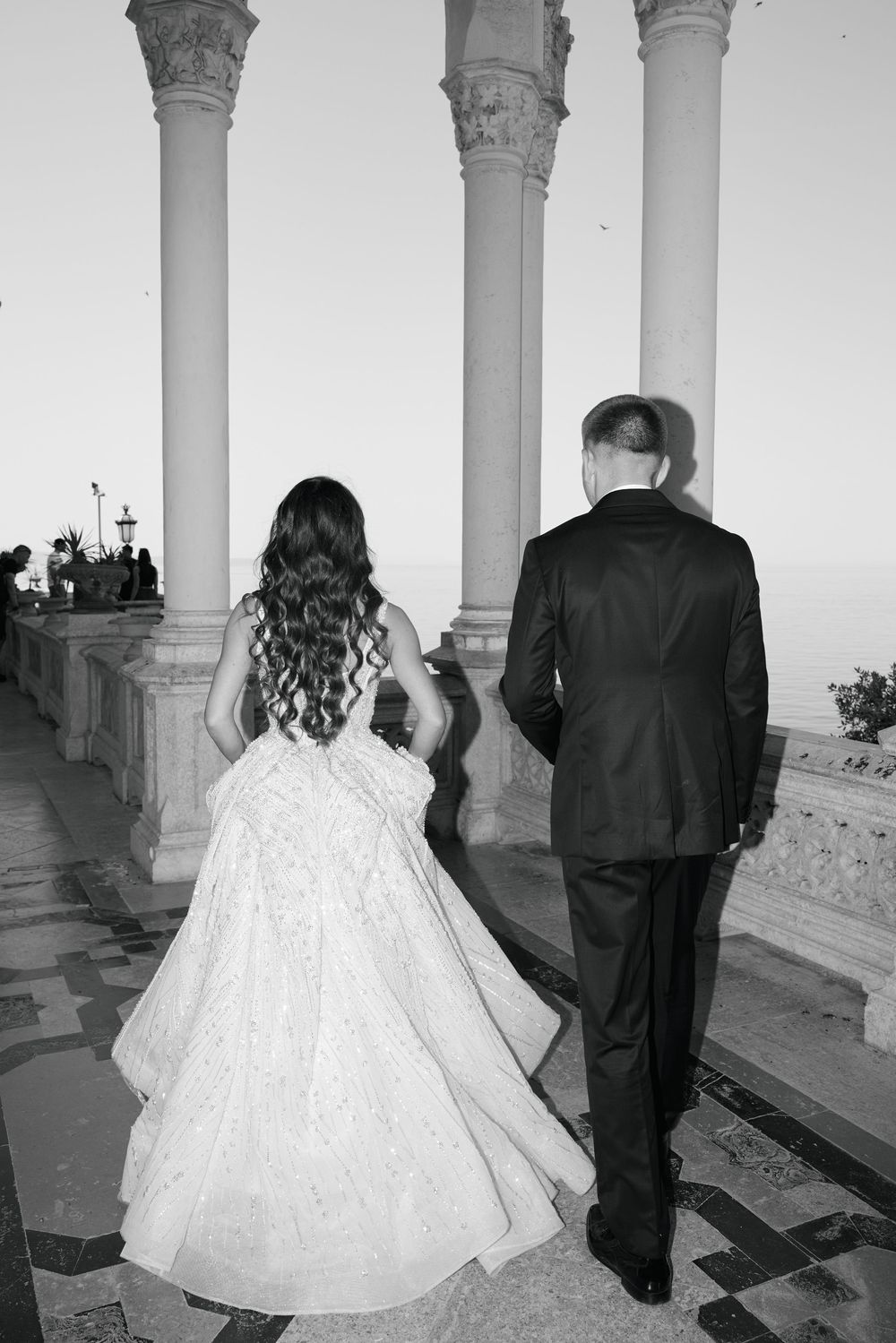 Bride and groom walk hand in hand between classical columns with ocean view at sunset wedding.
