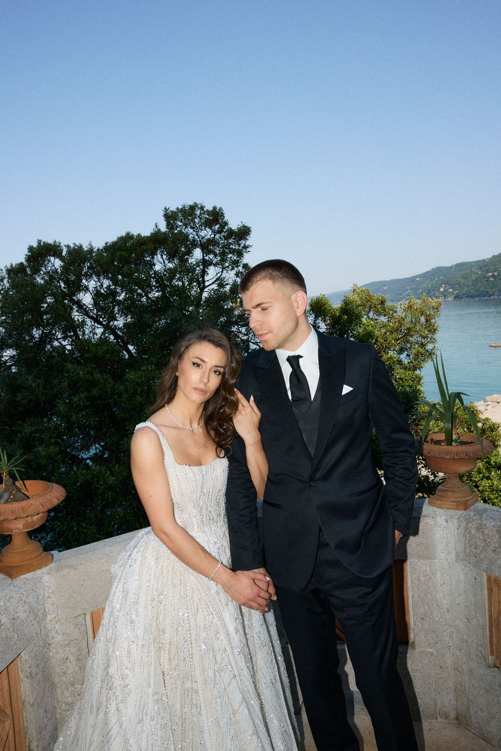 Bride and groom share romantic moment on terrace with ocean and greenery behind them.