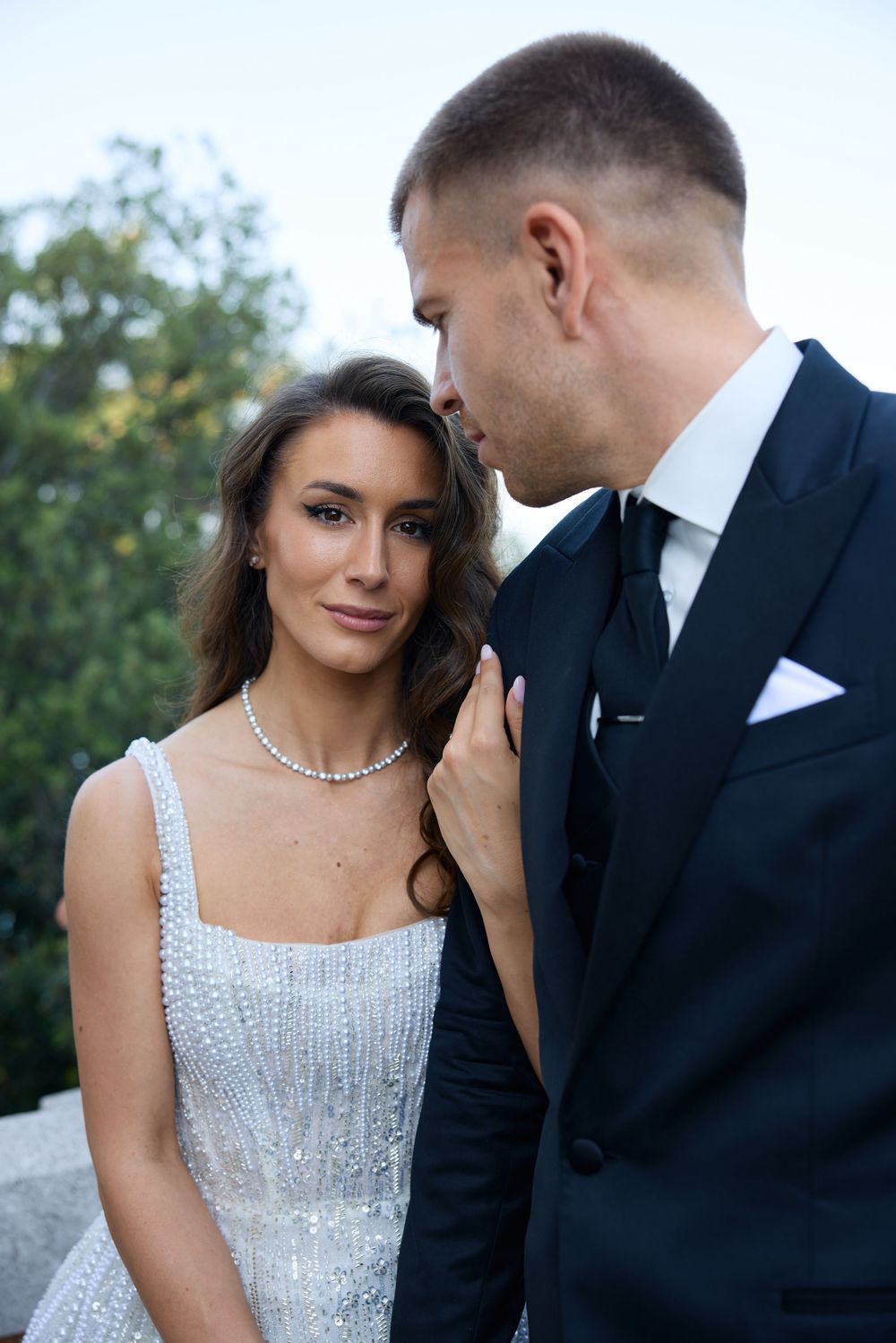 Bride in sparkling white gown with diamond necklace stands close to groom in black suit outdoors.
