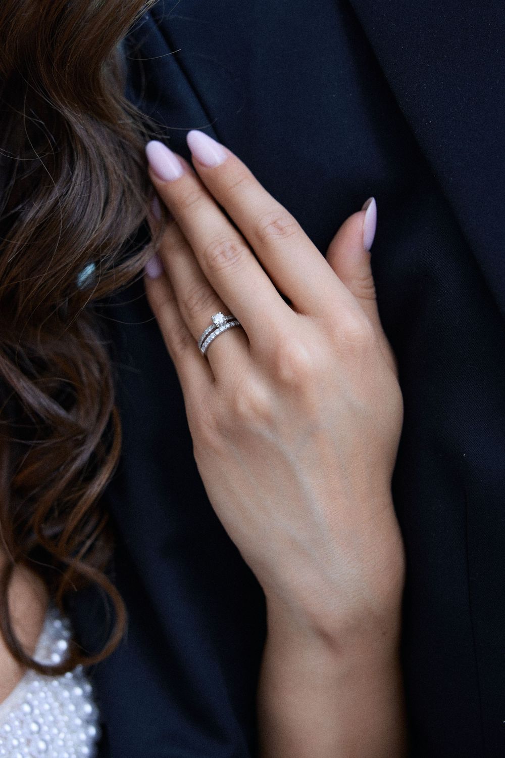 Close-up of a bride's hand with silver wedding rings and pink manicure resting on a groom's black suit jacket.