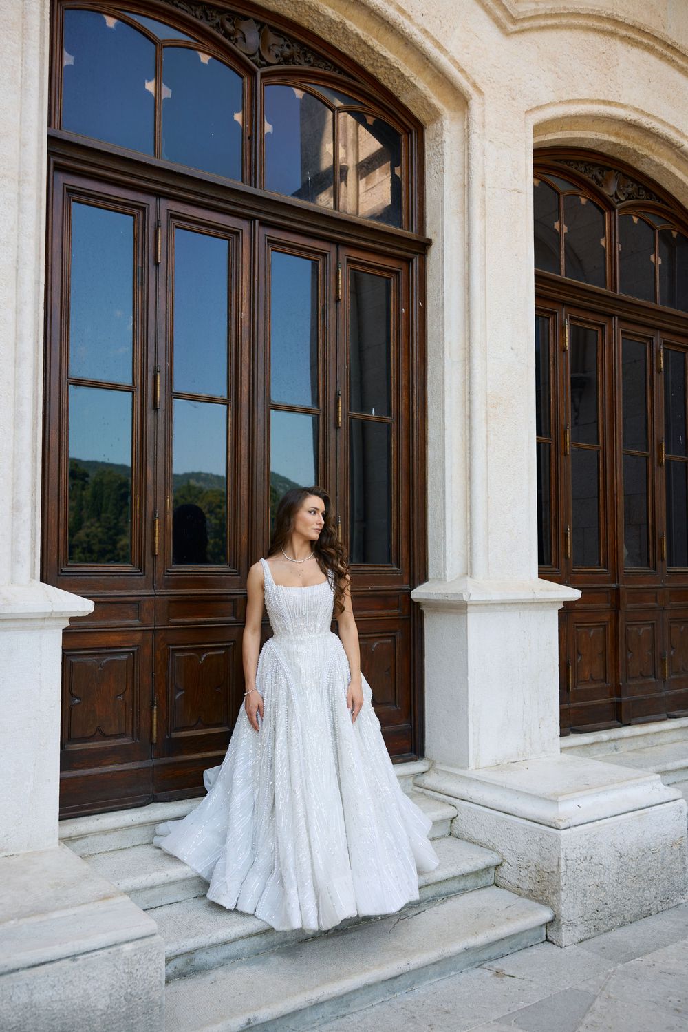 Bride in white lace ball gown wedding dress standing on steps of elegant building with ornate wooden doors.