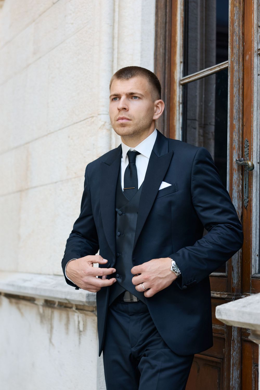 Groom in navy blue three-piece suit with black tie adjusting jacket outside historic stone building.