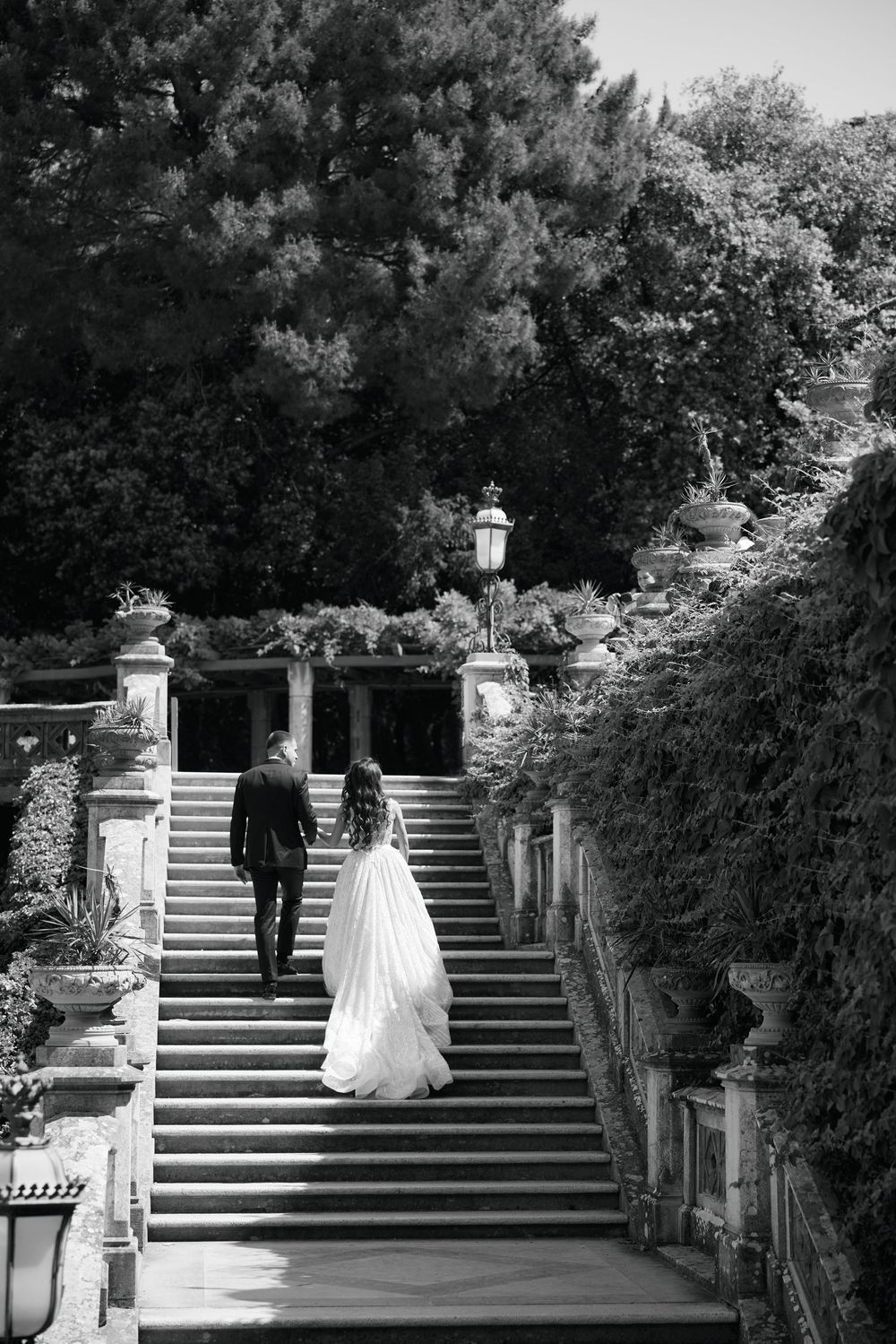 Bride and groom walk up ornate stone staircase surrounded by lush greenery in black and white wedding photo.