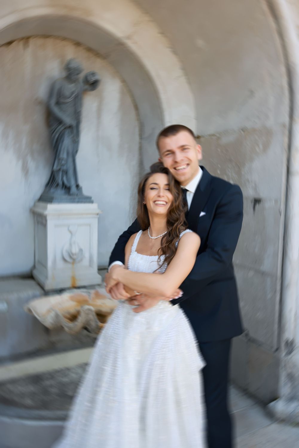 A joyful bride and groom embrace in front of a stone fountain with a classical statue during their wedding photo session.